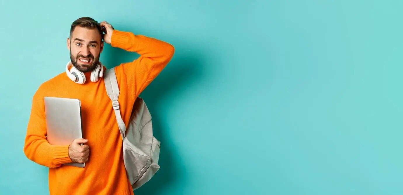 Ein verwirrter männlicher Student schaut ratlos, kratzt sich am Kopf und hält einen Laptop sowie einen Rucksack, während er steht.